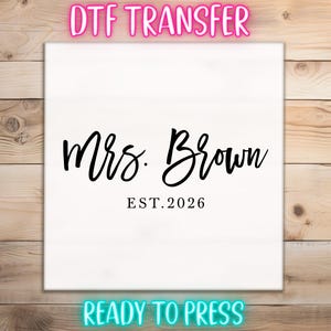 May include: White DTF transfer with the text "Mrs. Brown EST. 2026" in black script. The words "DTF TRANSFER" and "READY TO PRESS" are in neon pink and blue, respectively. The design is on a white background.