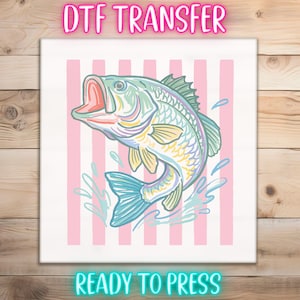 May include: DTF TRANSFER graphic featuring a colorful illustration of a leaping fish with an open mouth, set against a backdrop of pink vertical stripes. The words "DTF TRANSFER" and "READY TO PRESS" are displayed in neon-style text.