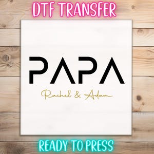 May include: White DTF transfer with the word "PAPA" in black block letters, with "Rachel & Adam" in gold script below. The words "DTF TRANSFER" and "READY TO PRESS" are in neon pink and blue, respectively.