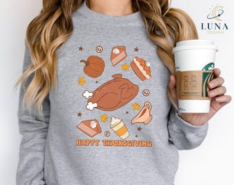 May include: A gray sweatshirt with a Thanksgiving-themed graphic. The design includes a roasted turkey, pumpkin, pie slices, and the text "HAPPY THANKSGIVING" in orange. A white coffee cup is held in the hand.