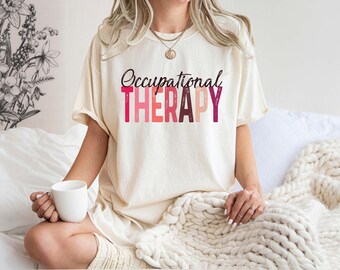 May include: Cream-colored t-shirt with the words "Occupational THERAPY" in a mix of pink and burgundy lettering. The person is holding a white mug. The shirt is part of a casual wear collection.