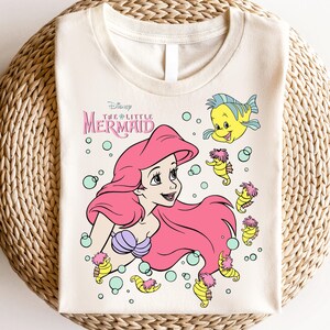 Retro vintage Ariel The Little Mermaid shirt featuring a nostalgic design with Ariel and Flounder. Made from soft, comfortable fabric for all-day wear. Perfect for Disney fans, princess lovers, and classic animation enthusiasts. Great for theme park visits, casual outfits, or as a unique gift. Available in multiple sizes and colors to match any style. A stylish and fun way to showcase love for this beloved Disney character.