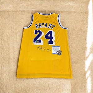Może przedstawiać: Żółta koszulka do koszykówki z napisem "BRYANT" i numerem "24" w kolorze niebieskim. Koszulka ma białe i niebieskie wykończenia i wydaje się być podpisana. Zawiera certyfikat autentyczności.
