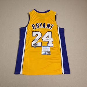 Może przedstawiać: Żółta koszulka do koszykówki z napisem "BRYANT" i numerem "24" w kolorze białym. Koszulka ma niebieskie i białe akcenty po bokach. Zawiera podpis i certyfikat autentyczności.