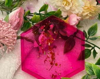 May include: A hexagonal pink resin coaster with gold flakes and dried flowers embedded inside. The coaster is sitting on a white fluffy surface.