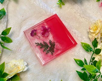 May include: A square pink and white resin tray with a clear border. The tray has a gradient of pink to white and contains pressed flowers and greenery.