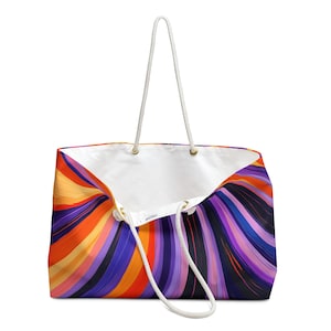 May include: A large tote bag with a vibrant, abstract design featuring swirling bands of orange, purple, and blue. The bag has white rope handles and a white interior. Gold-colored grommets secure the handles.