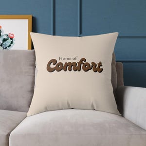 Peut inclure: Un coussin carré de couleur beige clair avec le texte "Home of Comfort" imprimé en brun foncé dans une police grasse et arrondie au centre. Le coussin présente une finition en tissu uni et lisse avec des bords cousus nets et précis.