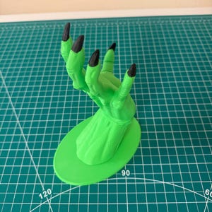 May include: A green witch's hand with black fingernails, reaching up, mounted on a green oval base.