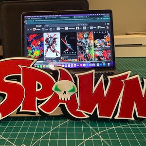 May include: A three-dimensional sign featuring the word SPAWN in bold red lettering with white and black outlines. A white skull icon with glowing green eyes replaces the letter A. The sign is positioned in front of an open laptop displaying images.