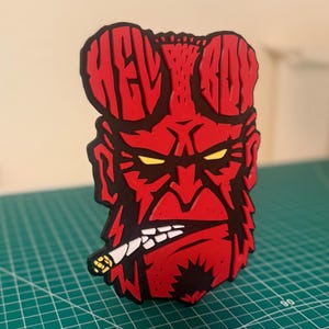 May include: Red 3D printed figurine of Hellboy, a comic book character, with the words "Hellboy" in red letters on his head. He is smoking a cigarette and has a serious expression on his face.