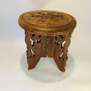 May include: A small, round, brown wooden stool featuring intricate floral carvings on the top surface. The base consists of two interlocking, foldable panels with openwork cutouts, showcasing a natural wood grain texture throughout the piece.