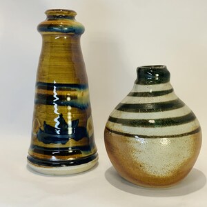May include: Two ceramic vases. The taller vase is cylindrical with a flared base, featuring horizontal bands of brown, tan, and deep blue glaze. The shorter, rounded vase has a cream-colored body with dark green horizontal stripes and a brown base.