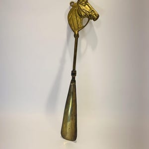 May include: A polished brass shoehorn featuring a detailed horse head handle at the top. The long, slender neck connects the horse head to a smooth, curved scoop designed for sliding into footwear. The metal has a warm, golden metallic finish.