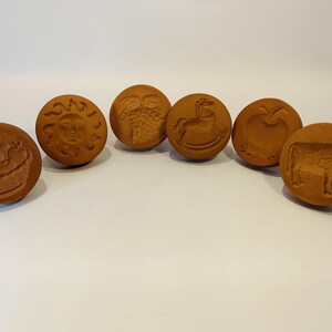 May include: A set of six circular brown clay stamps, each featuring a distinct embossed design including a basket, a sun with a face, a cluster of grapes, a rocking horse, an apple, and a cow, arranged in a semi-circular formation.