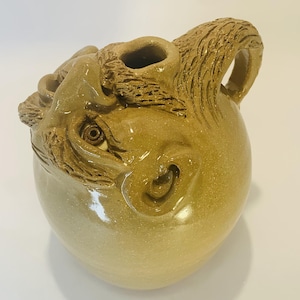May include: A tan ceramic jug featuring a sculpted face on its upper surface. The vessel has a smooth, glossy finish, a single handle, and a prominent eye and ear integrated into the clay form near the top opening of the container.