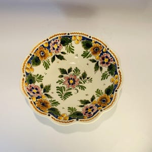 May include: A round, off-white ceramic plate with a scalloped edge. The plate is decorated with a floral pattern in shades of yellow, pink, blue, and green. The rim has a dark blue border with a gold trim.