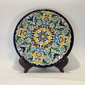 May include: A decorative ceramic plate with a dark blue rim and intricate floral patterns in yellow, blue, green, and red. The plate is displayed on a dark brown wooden stand, showcasing its detailed design and vibrant colors.
