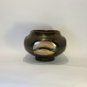 May include: A handcrafted ceramic vase with a dark brown glaze. The vase has a rounded body, a small foot, and a wide opening. A lighter-colored, oval-shaped design is on the front. The vase is suitable for displaying flowers or as a decorative item.