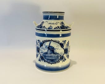 May include: A ceramic milk can with a cream-colored base and blue hand-painted floral patterns. The front features a central oval illustration of a windmill with the word Holland written above it. It has two small side handles and a lid.