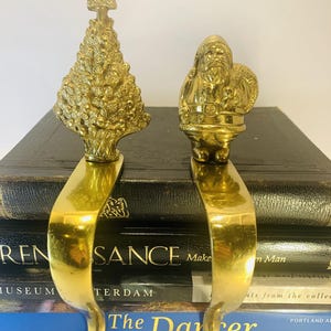 May include: Two polished brass stocking holders featuring a textured Christmas tree with a star topper and a detailed Santa Claus figure. Each ornament is attached to a smooth, curved metal hook designed for hanging holiday stockings.