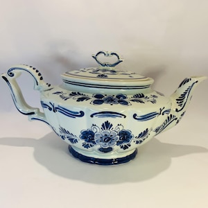 May include: A white ceramic teapot featuring intricate blue floral patterns and scrollwork designs. The teapot includes a matching lid with a decorative handle, a curved spout, and a shaped handle, all decorated with hand-painted blue motifs.