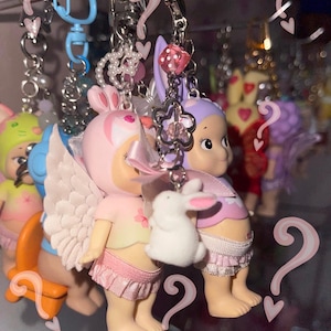 May include: A collection of colorful keychains featuring small figurines. The keychains include angel-winged figures in pink and purple outfits, each holding a small white bunny. Other keychains have various charms and beads.