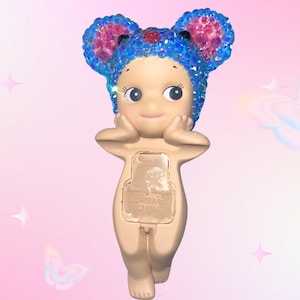 Puede incluir: Una figura caprichosa de una peque&ntilde;a figura desnuda con grandes orejas azules brillantes y un tocado a juego. La figura tiene una expresi&oacute;n serena, con las manos tocando suavemente sus mejillas. La parte posterior de la figura tiene un panel rectangular con texto.