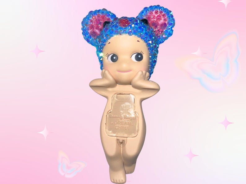 Puede incluir: Una figura caprichosa de una peque&ntilde;a figura desnuda con grandes orejas azules brillantes y un tocado a juego. La figura tiene una expresi&oacute;n serena, con las manos tocando suavemente sus mejillas. La parte posterior de la figura tiene un panel rectangular con texto.