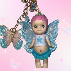 May include: A whimsical charm featuring a doll with blue fairy wings, a strawberry hat, and a white t-shirt with "Cafe Time" printed on it. A gold chain holds a butterfly charm with blue and pink accents. The background is pink.