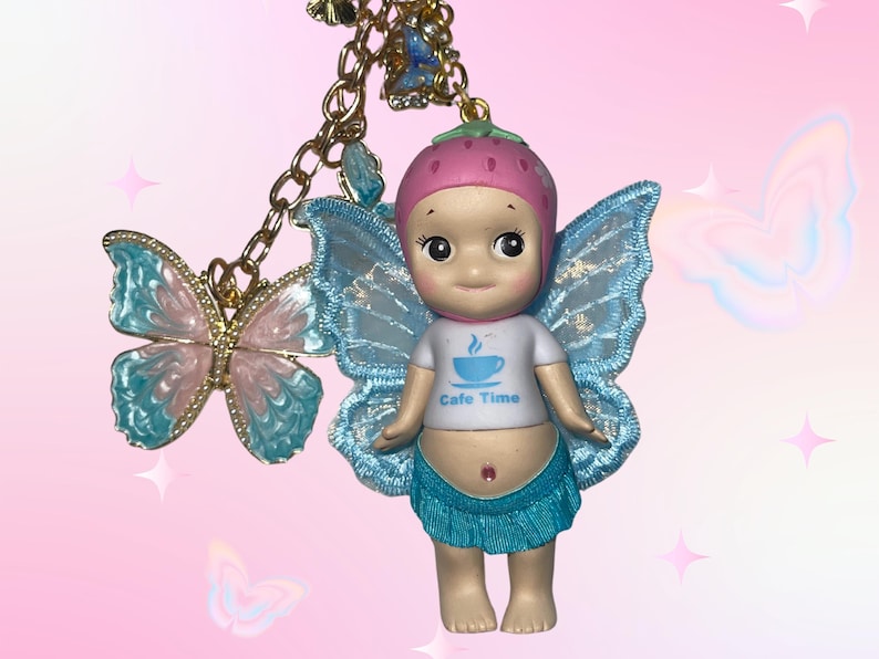 May include: A whimsical charm featuring a doll with blue fairy wings, a strawberry hat, and a white t-shirt with "Cafe Time" printed on it. A gold chain holds a butterfly charm with blue and pink accents. The background is pink.