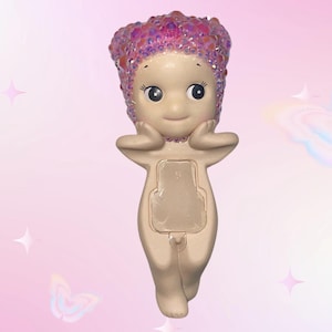 Puede incluir: Una figura caprichosa con un cuerpo beige claro, ojos grandes y una expresi&oacute;n sonriente. La figura tiene una cubierta para la cabeza rosa brillante y posa con las manos cerca de la cara. La figura tiene una abertura rectangular en la parte delantera.