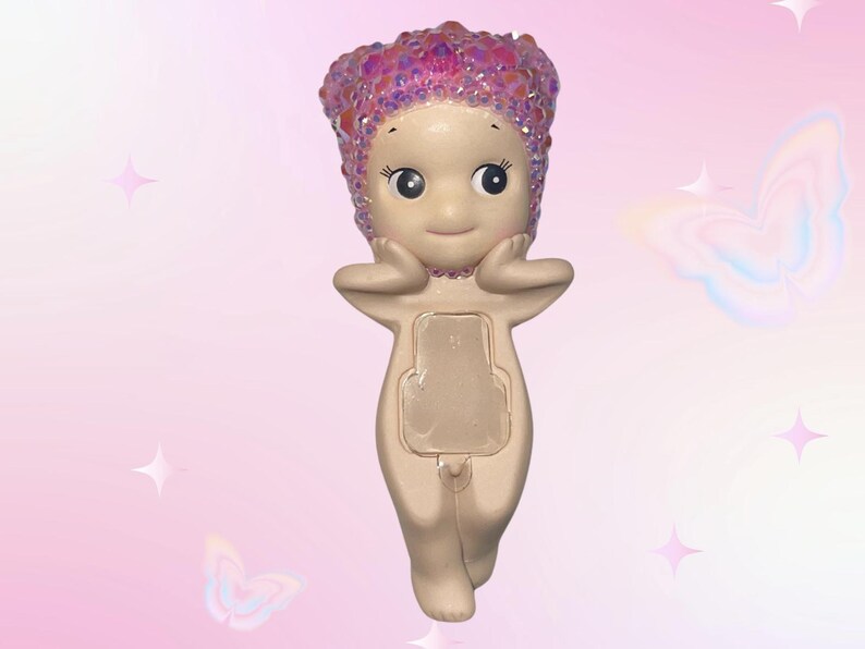 Puede incluir: Una figura caprichosa con un cuerpo beige claro, ojos grandes y una expresi&oacute;n sonriente. La figura tiene una cubierta para la cabeza rosa brillante y posa con las manos cerca de la cara. La figura tiene una abertura rectangular en la parte delantera.