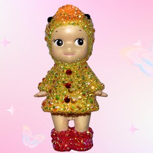 Puede incluir: Figura decorativa de un personaje parecido a una mu&ntilde;eca con un atuendo amarillo y naranja enjoyado. La figura tiene una cara sonriente, botas rojas enjoyadas y un sombrero a juego enjoyado.