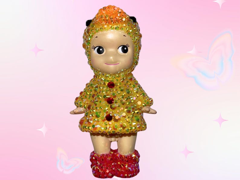 Puede incluir: Figura decorativa de un personaje parecido a una mu&ntilde;eca con un atuendo amarillo y naranja enjoyado. La figura tiene una cara sonriente, botas rojas enjoyadas y un sombrero a juego enjoyado.