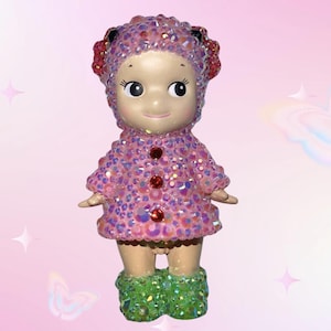 Puede incluir: Figura decorativa de mu&ntilde;eca con abrigo y sombrero rosas, ambos cubiertos de pedrer&iacute;a iridiscente. La mu&ntilde;eca tiene una cara sonriente, detalles rojos y botas verdes brillantes. Fondo rosa suave con estrellas y mariposas.