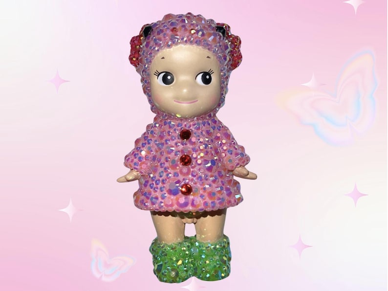 Puede incluir: Figura decorativa de mu&ntilde;eca con abrigo y sombrero rosas, ambos cubiertos de pedrer&iacute;a iridiscente. La mu&ntilde;eca tiene una cara sonriente, detalles rojos y botas verdes brillantes. Fondo rosa suave con estrellas y mariposas.