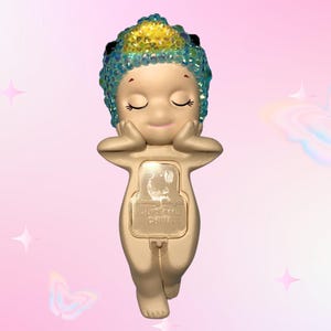 May include: A small, beige figurine of a cherubic figure with closed eyes, hands supporting its face. The figure has a jeweled headpiece with blue and yellow accents. The base of the figurine has a rectangular panel with text. The background is pink with butterfly and star graphics.