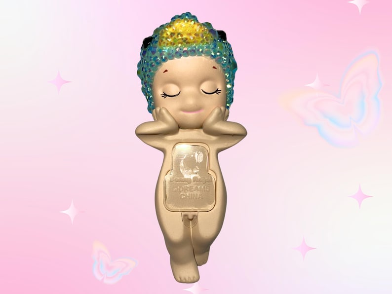 May include: A small, beige figurine of a cherubic figure with closed eyes, hands supporting its face. The figure has a jeweled headpiece with blue and yellow accents. The base of the figurine has a rectangular panel with text. The background is pink with butterfly and star graphics.