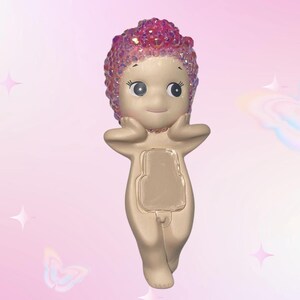 Puede incluir: Una figura caprichosa de una figura desnuda con una cara sonriente y una cabeza rosa cubierta de cristales. La figura se est&aacute; sosteniendo las manos en la cara. El fondo es un degradado de rosa con gr&aacute;ficos de estrellas y mariposas.