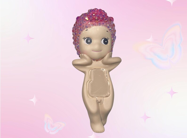 Puede incluir: Una figura caprichosa de una figura desnuda con una cara sonriente y una cabeza rosa cubierta de cristales. La figura se est&aacute; sosteniendo las manos en la cara. El fondo es un degradado de rosa con gr&aacute;ficos de estrellas y mariposas.