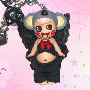 May include: A keyring featuring a small doll dressed in a grey koala hood with red spiral cheek markings and red eyes. The doll wears a black top with a red bow, black trousers, and black lace wings. A silver chain with black dice is attached.