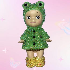 Puede incluir: Figura decorativa de un beb&eacute; vestido con un disfraz de rana verde. El disfraz est&aacute; cubierto de adornos brillantes verdes y transparentes. La figura tiene una cara sonriente y botas amarillas.