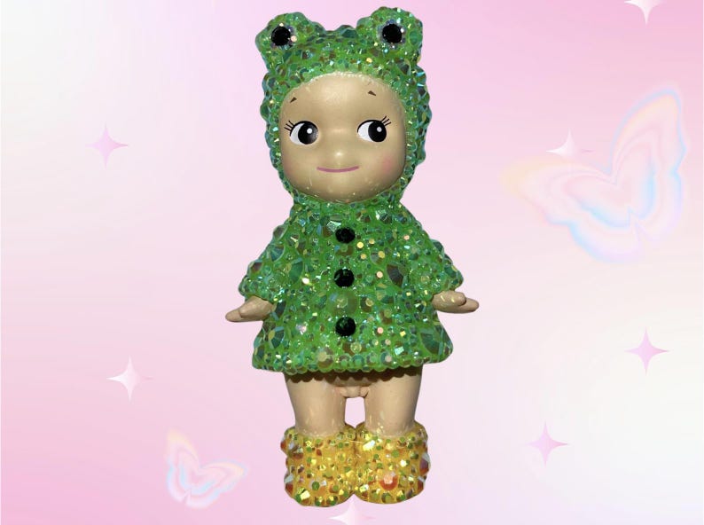 Puede incluir: Figura decorativa de un beb&eacute; vestido con un disfraz de rana verde. El disfraz est&aacute; cubierto de adornos brillantes verdes y transparentes. La figura tiene una cara sonriente y botas amarillas.