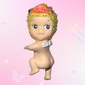 May include: A small, nude figurine with a smiling face and large eyes. The figurine has a head covering with orange, yellow, and gold glitter. Small white wings are attached to the back. The background is pink with butterfly and star graphics.