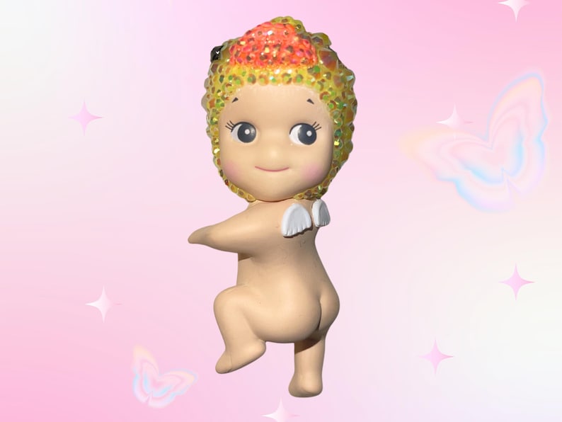 May include: A small, nude figurine with a smiling face and large eyes. The figurine has a head covering with orange, yellow, and gold glitter. Small white wings are attached to the back. The background is pink with butterfly and star graphics.