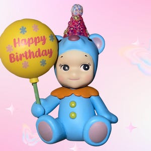 Puede incluir: Una figura de oso azul que lleva un sombrero de fiesta rosa con lentejuelas y un cuello naranja. El oso tiene un traje azul claro con dos botones verdes y sostiene un globo amarillo con el texto Happy Birthday e iconos de flores impresos.