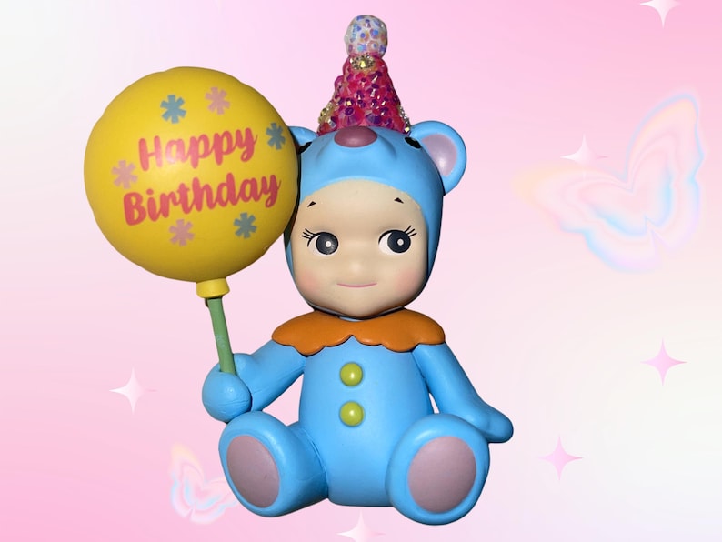 Puede incluir: Una figura de oso azul que lleva un sombrero de fiesta rosa con lentejuelas y un cuello naranja. El oso tiene un traje azul claro con dos botones verdes y sostiene un globo amarillo con el texto Happy Birthday e iconos de flores impresos.