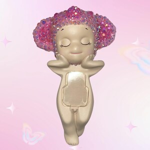 Puede incluir: Una figura caprichosa de una figura serena con los ojos cerrados, las manos sosteniendo suavemente la cara. La figura tiene un cuerpo beige claro y una cabeza de purpurina rosa brillante. La figura tiene forma rectangular en la parte delantera.