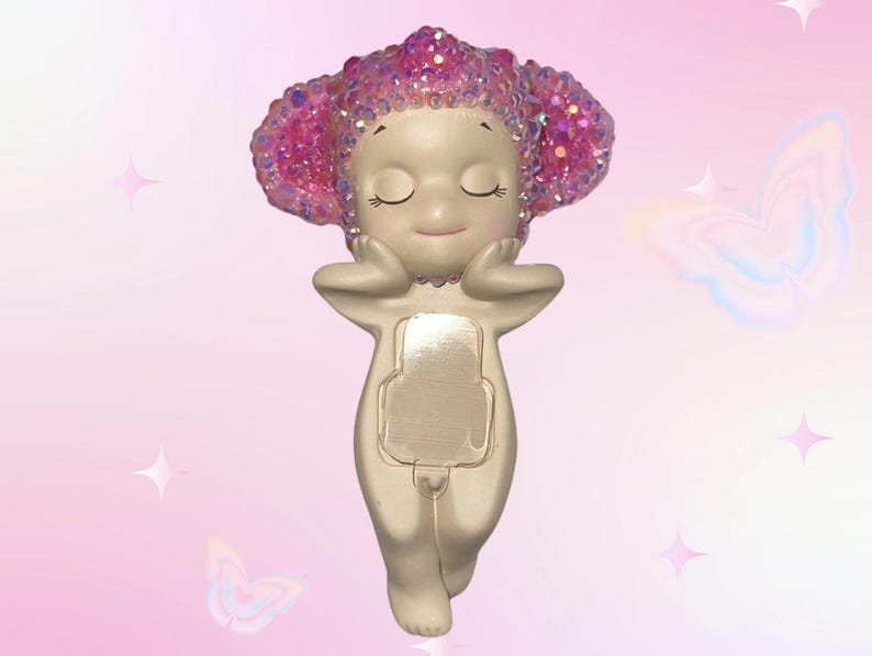 Puede incluir: Una figura caprichosa de una figura serena con los ojos cerrados, las manos sosteniendo suavemente la cara. La figura tiene un cuerpo beige claro y una cabeza de purpurina rosa brillante. La figura tiene forma rectangular en la parte delantera.
